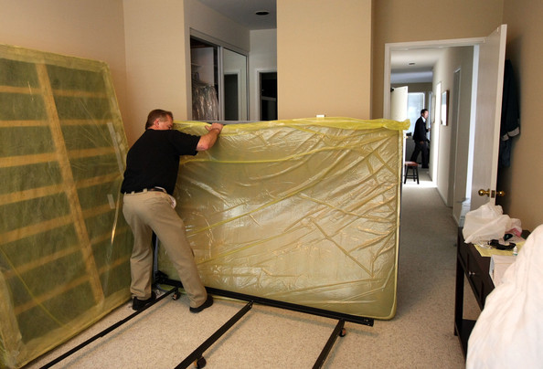 A pest control worker in a black shirt and khaki pants is moving a mattress sealed in yellow plastic wrap as part of a bed bug treatment protocol.